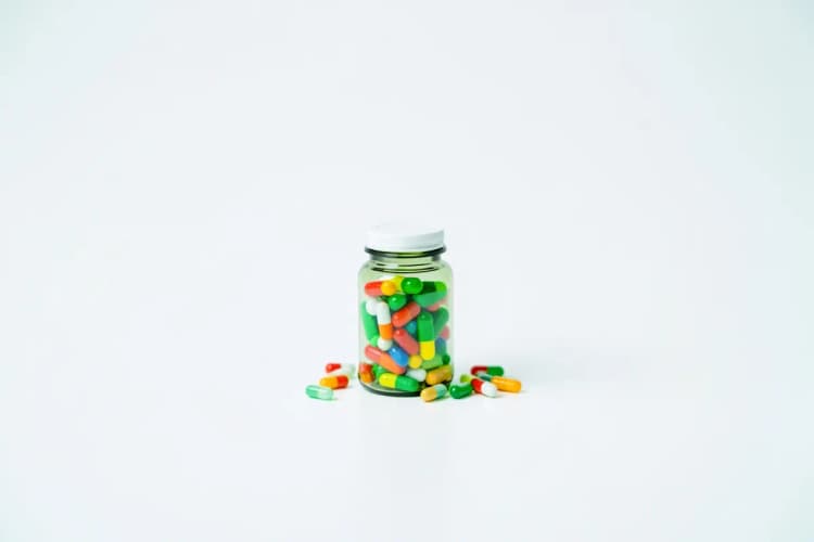 Frasco de suplemento vitamínico com cápsulas coloridas ao redor, sobre fundo branco.