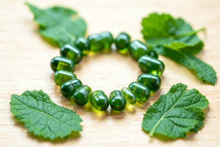 Cápsulas de colágeno dispostas em forma de círculo sobre uma superfície de madeira clara, com algumas folhas verdes espalhadas ao redor.