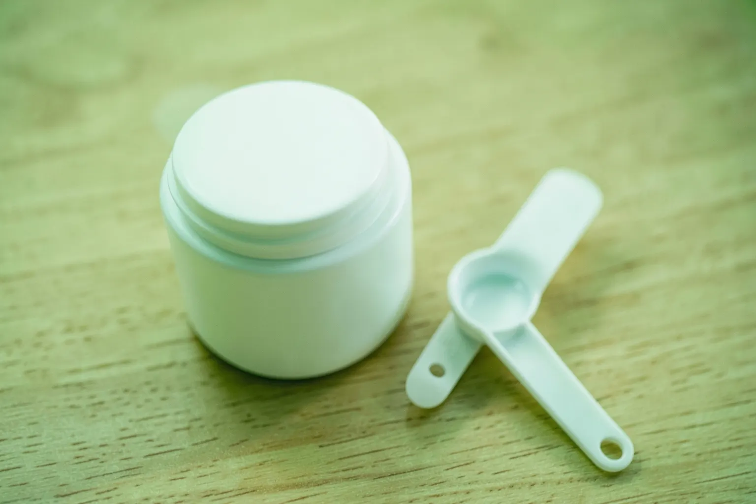 Pote de colágeno em pó com uma colher medidora ao lado, sobre uma superfície de madeira clara.