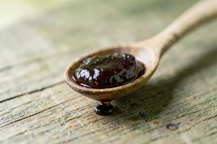 Uma colher de chá cheia de própolis marrom escura, com algumas gotas caindo sobre uma superfície de madeira.