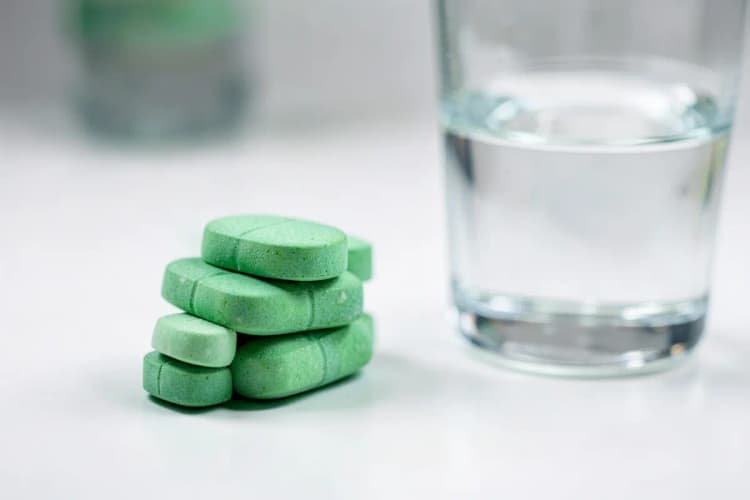 Uma pilha de comprimidos de vitamina C ao lado de um copo de água em uma superfície branca limpa.