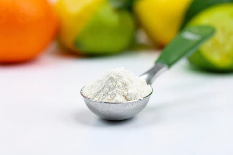 Um medidor de colheres com pó de vitamina C em um fundo branco, com algumas frutas cítricas desfocadas ao fundo.