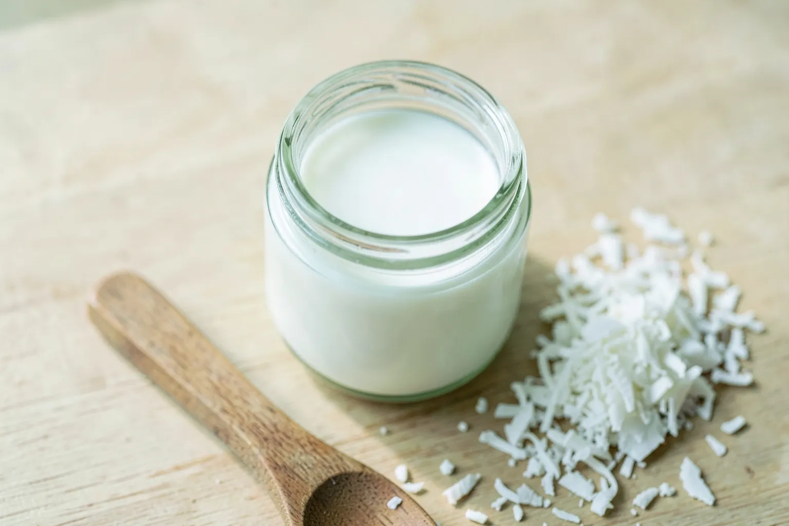 Um pote de vidro com óleo de coco ao lado de uma colher e coco ralado sobre uma superfície de madeira clara.