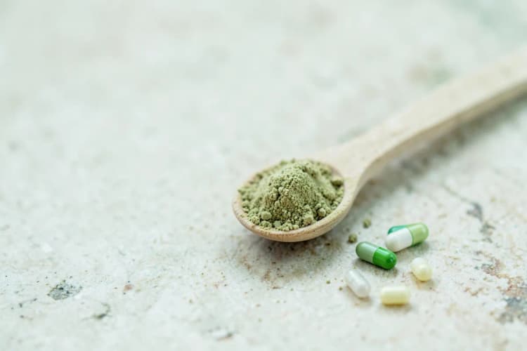 Uma colher de chá com maca peruana em pó e algumas cápsulas ao lado em uma superfície de pedra clara.