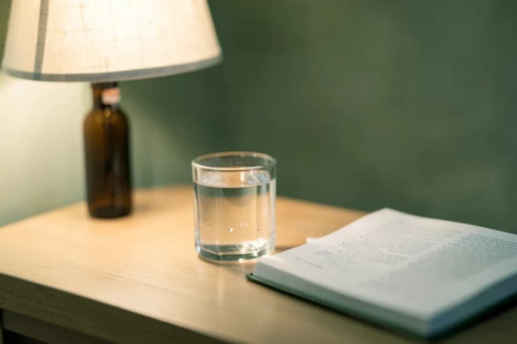 Um frasco de melatonina em gotas ao lado de um copo com água e um livro aberto sobre uma mesa de cabeceira.