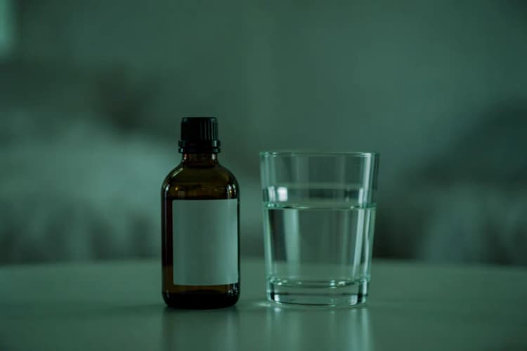 Um frasco de melatonina em gotas e um copo de água sobre uma mesa com um fundo desfocado de um quarto.