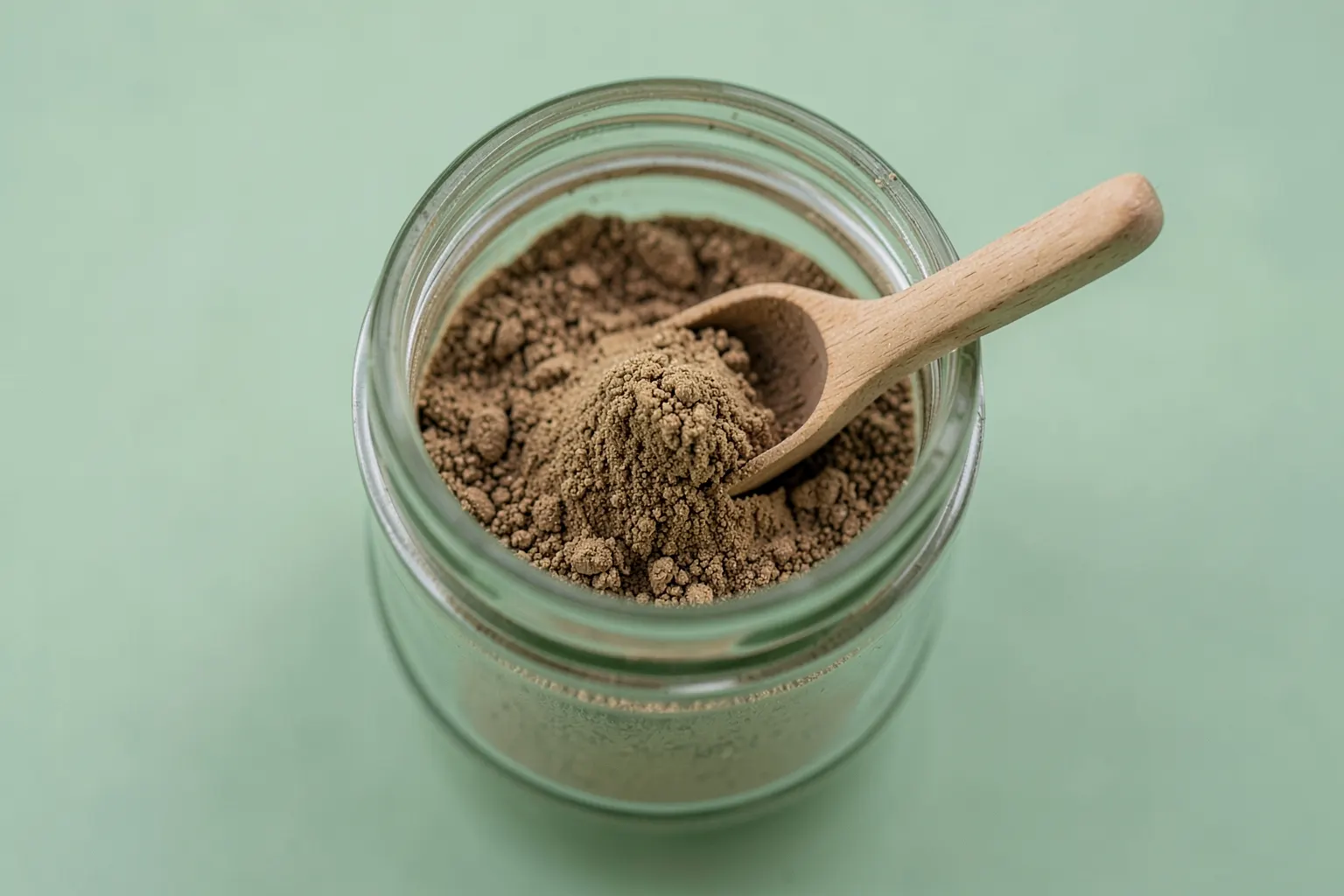 Tribulus terrestris em pó em um recipiente de vidro com uma colher ao lado em um fundo de madeira clara.