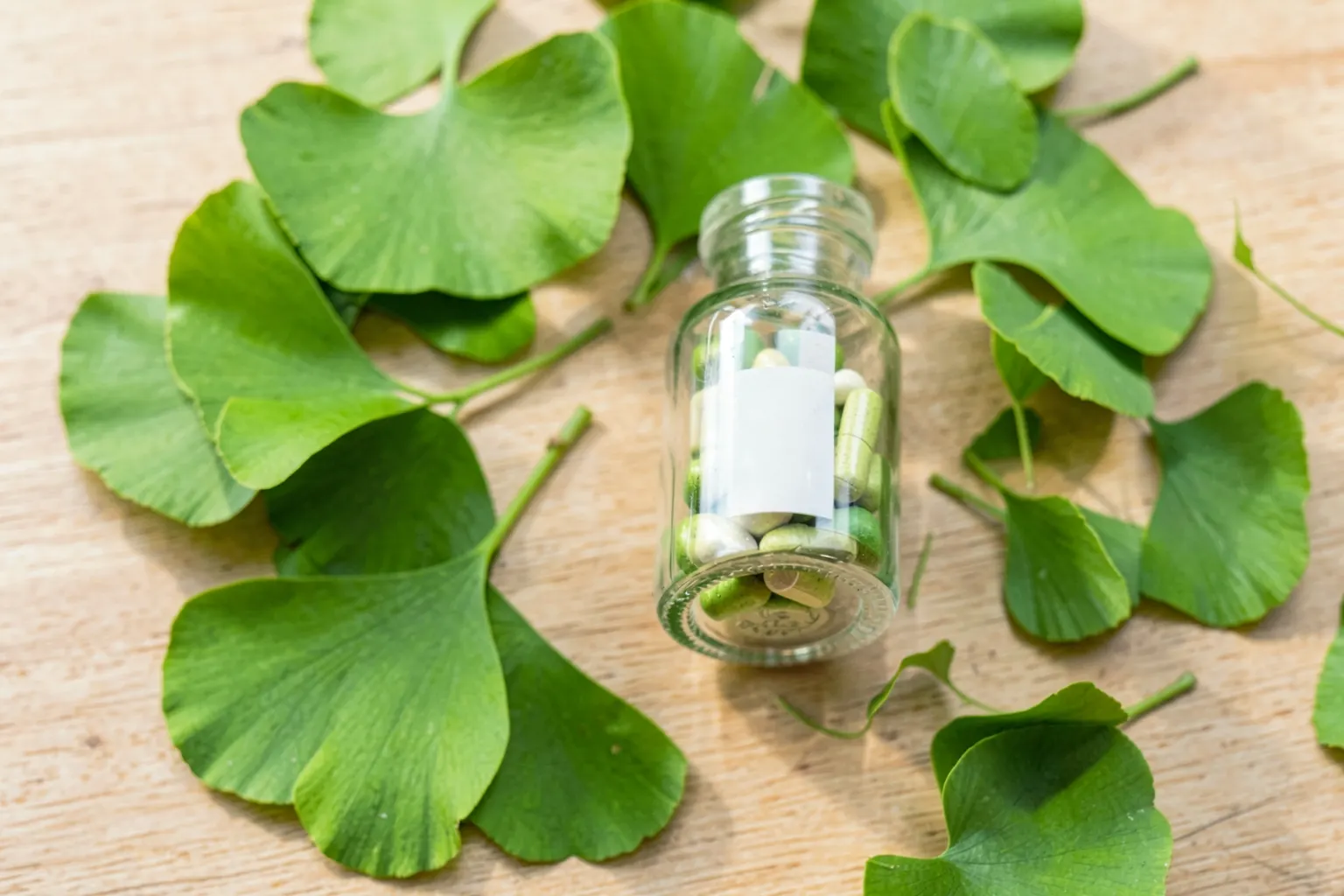 Um pote de comprimidos de Ginkgo Biloba ao lado de folhas verdes de Ginkgo Biloba sobre uma superfície de madeira clara.