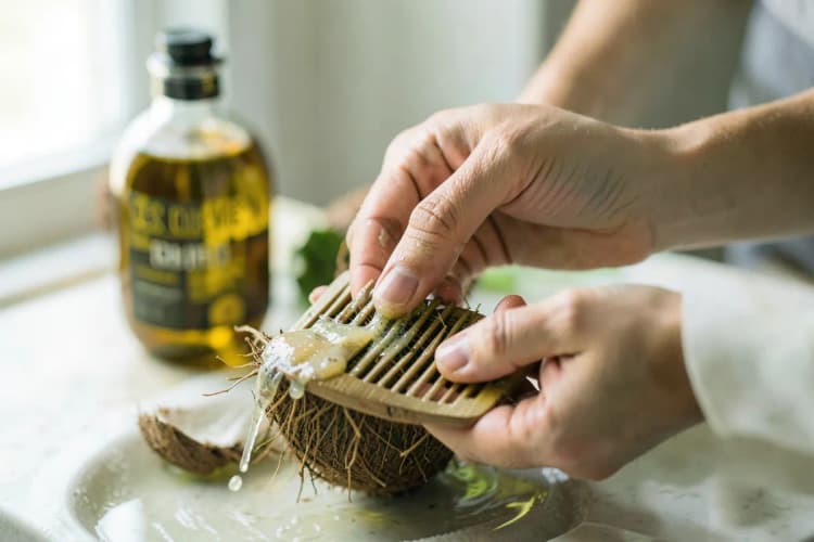 Mãos aplicando óleo de coco em um pente para cabelo, com um frasco de óleo e um coco ao fundo.