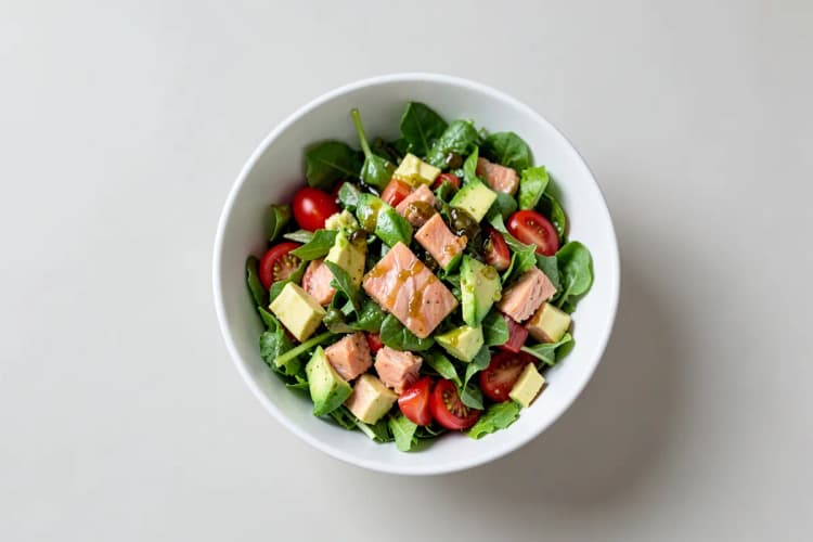 Uma tigela de salada com folhas verdes, tomate cereja, abacate e pedaços de salmão, com um fio de azeite.